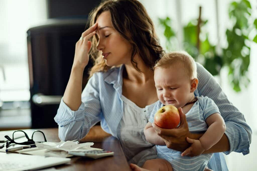 Overwhelmed mother holding baby at table illustrating parenting stress and modern parenting pressure