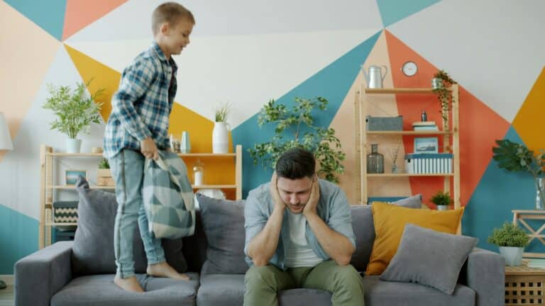 The Moment Parenting Became About Fear Instead of Values 5 Overwhelmed father sitting on couch with energetic child illustrating family stress and fear of child missing out