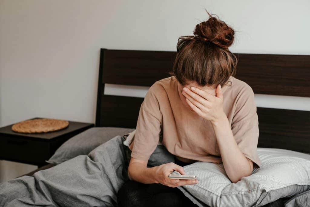 Phone addiction psychology: Woman sitting in bed stressed while looking at her phone illustrating social media addiction and mental fatigue